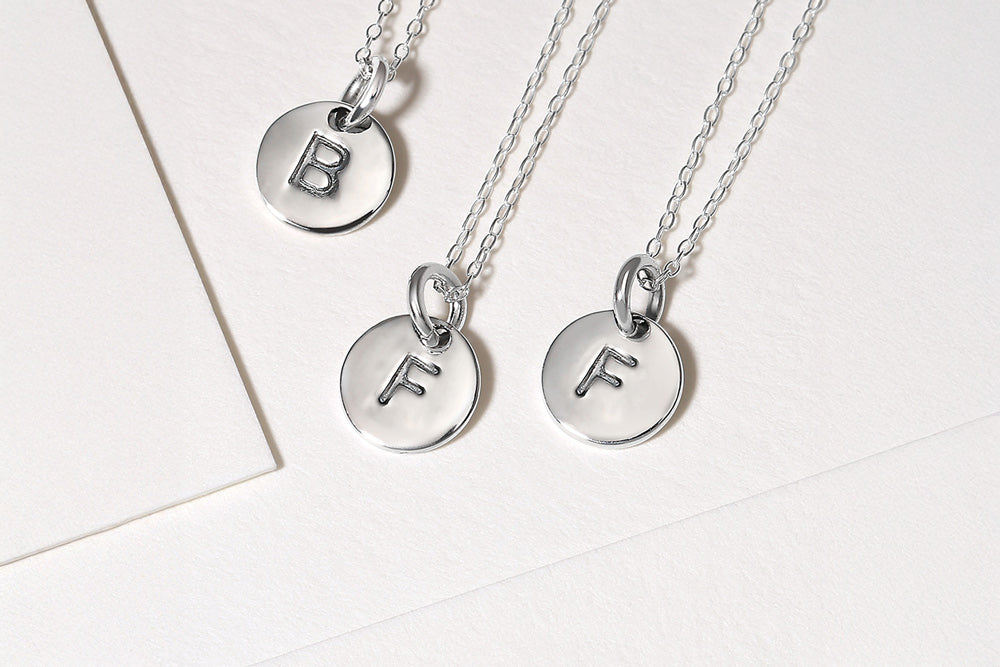 Three silver necklaces with round pendants engraved with initials on a light background.